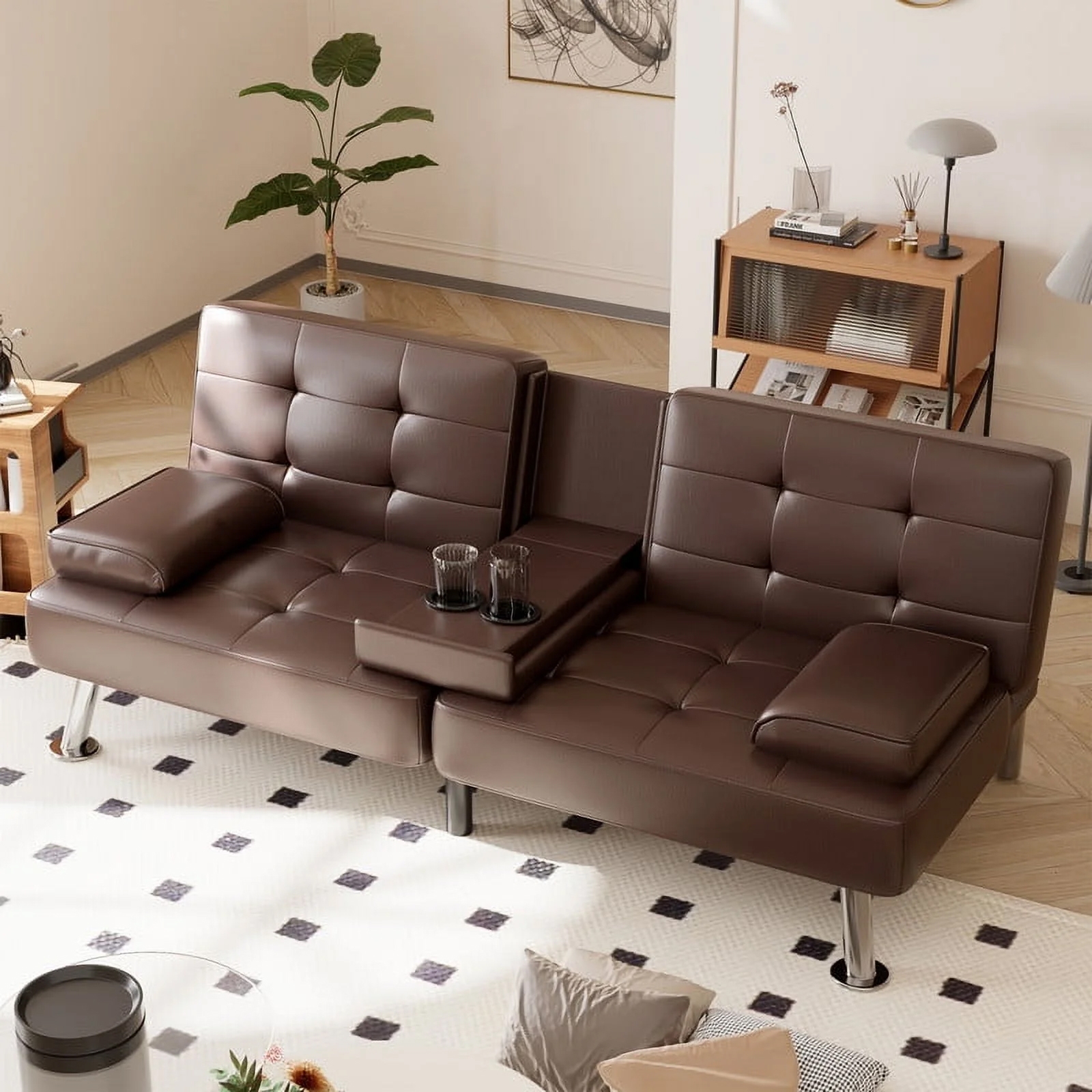 A modern brown leather sofa with a central armrest featuring built-in cup holders. The sofa is in a minimalist living room with a light-colored rug, decorative plants, and a wooden shelf in the background.