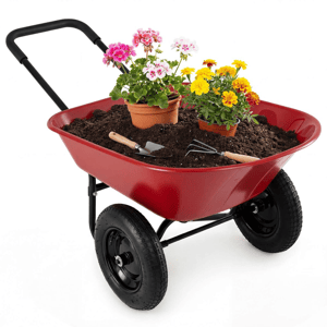 A red wheelbarrow filled with soil holds potted pink geraniums and yellow marigolds, with a small gardening trowel and hand rake.
