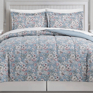 A floral-patterned bedding set with a comforter and two pillow shams on a bed with a white headboard. The design features pastel blue, pink, and white flowers.