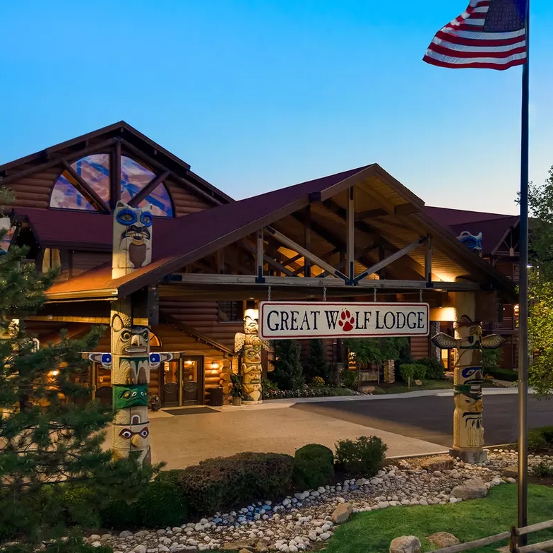 Entrance of Great Wolf Lodge featuring totem poles, a lit entrance, and an American flag, set against a background of trees and a clear sky.