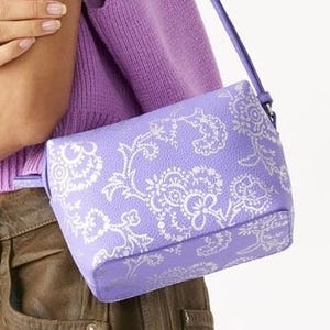 A small lavender bag with a white floral pattern, carried on the shoulder over a purple sweater, paired with brown pants.