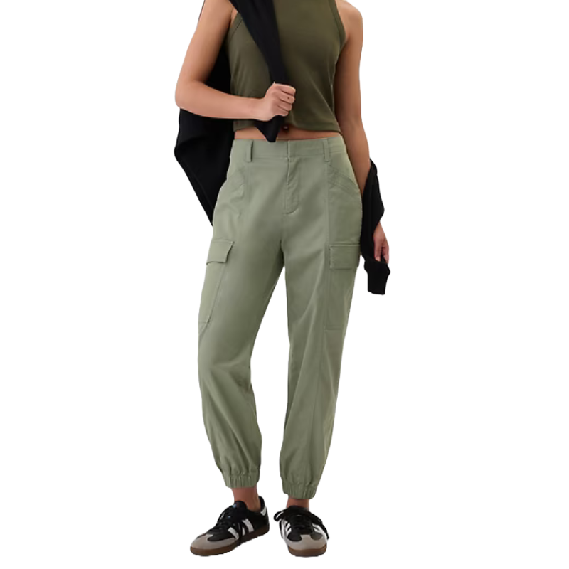 Olive green cargo joggers with elasticized cuffs, side pockets, and a relaxed fit. Paired with a cropped olive tank top, a black jacket draped over one shoulder, and black sneakers.