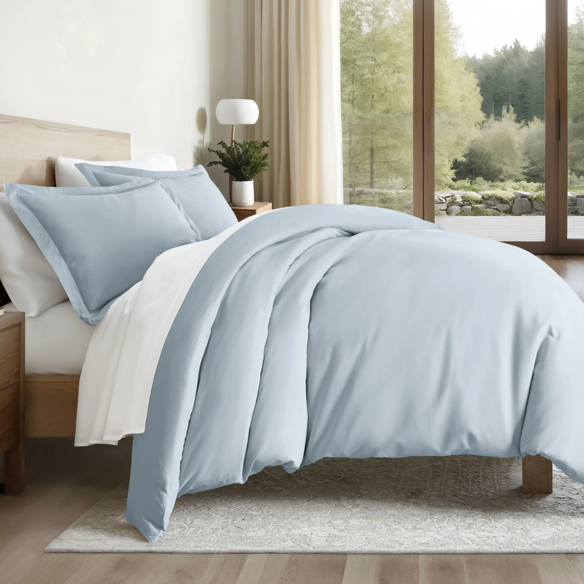 A bedroom features a bed with light blue and white bedding, including a duvet and pillows. The setup includes a wooden nightstand, a lamp, and small plants, with a large window revealing a forest view.
