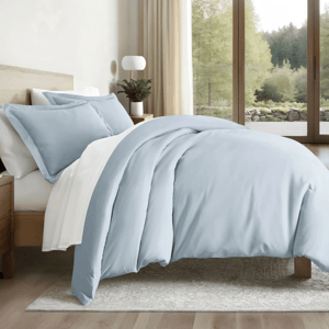 A bedroom features a bed with light blue and white bedding, including a duvet and pillows. The setup includes a wooden nightstand, a lamp, and small plants, with a large window revealing a forest view.