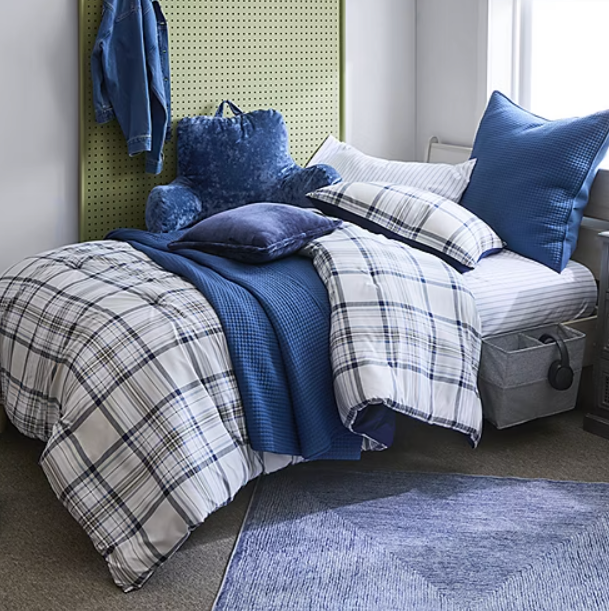 A cozy bedroom setup featuring a plaid-patterned comforter, blue pillows, and a plush backrest on a bed against a perforated green headboard, with additional storage in a gray basket and a blue area rug beneath.