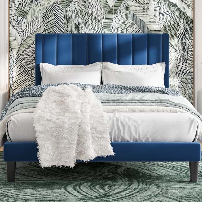 A blue upholstered bed with a vertically striped headboard is set against a leafy wallpaper. It features white and blue bedding with a fluffy white throw, complemented by a green circular-patterned rug.