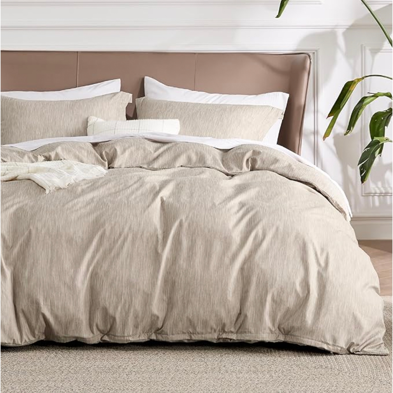A beige 3-piece Queen Duvet Cover Set includes a duvet cover and two pillow shams, featuring a subtle textured pattern. The set is displayed on a neatly made bed with a beige and white color scheme, accented by a plant in the background.