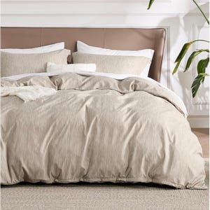 A beige 3-piece Queen Duvet Cover Set includes a duvet cover and two pillow shams, featuring a subtle textured pattern. The set is displayed on a neatly made bed with a beige and white color scheme, accented by a plant in the background.