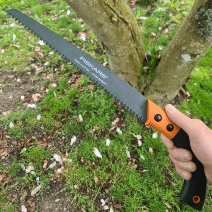 A hand holding a Fiskars saw with a black and orange handle, used for cutting wood.