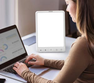 A woman is working on a laptop next to a light therapy lamp with touch controls.