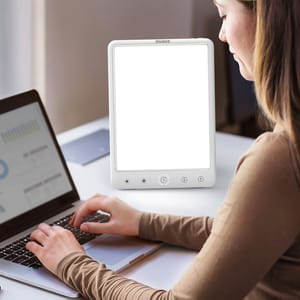 A woman is working on a laptop next to a light therapy lamp with touch controls.