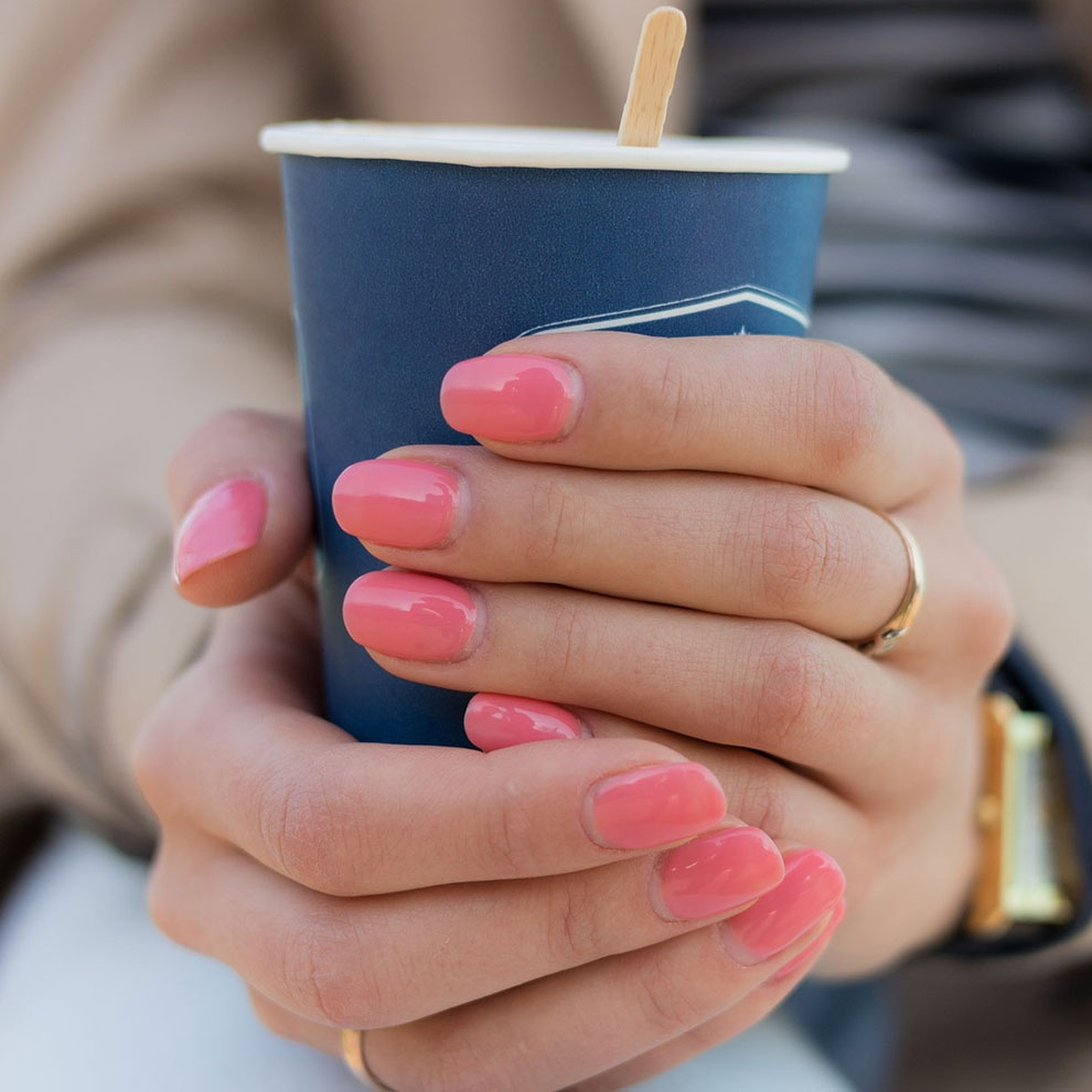 A person holds a blue cup while showing off their pink manicured nails and wearing a gold ring and wristwatch.