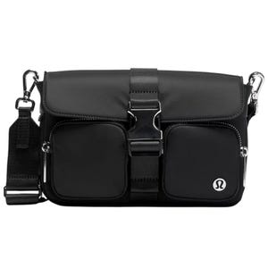 A black crossbody bag with two front pockets, adjustable strap, prominent buckle closure, and a logo displayed on the lower right corner.