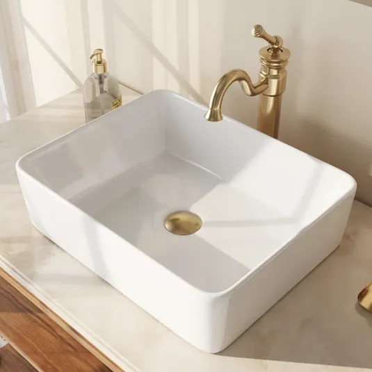 A rectangular white sink with a gold circular drain is set on a marble countertop. Nearby, a gold faucet and a clear soap dispenser with a pump are visible.