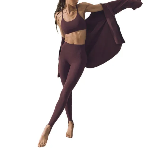 A woman is wearing high-rise stirrup leggings in a dark burgundy color. The leggings are paired with a matching sports bra, and she holds a matching jacket.