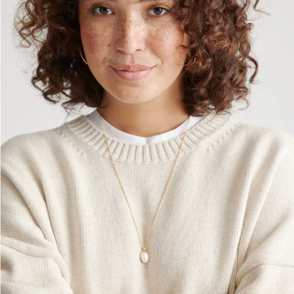 A person wearing a cream-colored knit sweater and a gold necklace with a small pearl pendant.