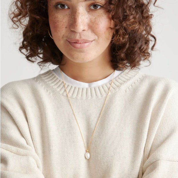 A person wearing a cream-colored knit sweater and a gold necklace with a small pearl pendant.