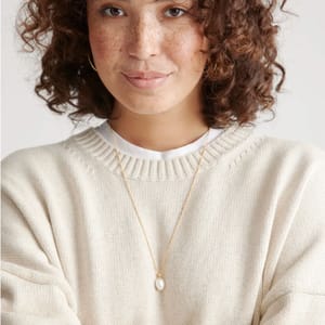 A person wearing a cream-colored knit sweater and a gold necklace with a small pearl pendant.