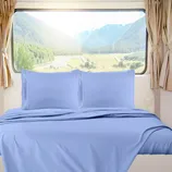 A bed with light blue sheets and pillowcases is placed against a large window overlooking a mountainous landscape. Light beige curtains frame the window.