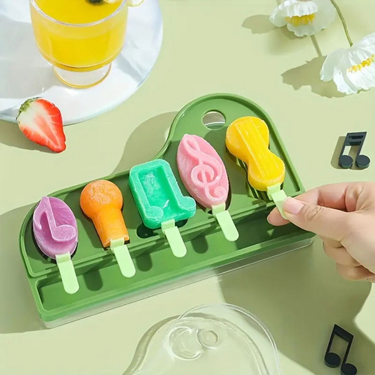 The Piano Key Ice Cube & Popsicle Mold Set features five musical-themed molds, including shapes like a musical note and treble clef. The popsicles come in various colors, displayed on a green tray.