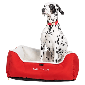 A Dalmatian sits on a red Kate Spade dog bed with white fleece lining and the phrase \“CALL IT A DAY\“ printed on the front.