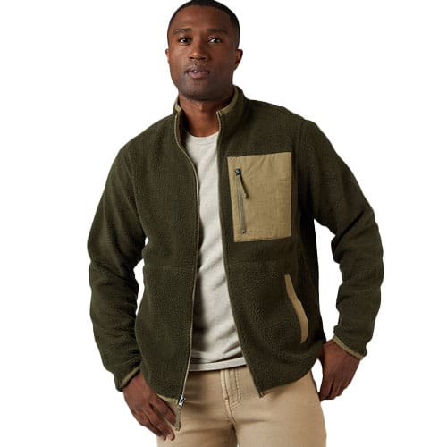 A man wears an olive green fleece jacket with khaki trim and zippered chest and side pockets, paired with a light beige shirt and pants.