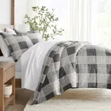 A bed with a black and gray plaid comforter set, including matching pillow shams, is shown in a bright, minimalistic bedroom with a wooden side table and a potted plant for decoration.