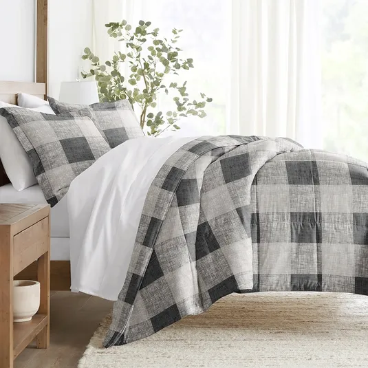 A bed with a black and gray plaid comforter set, including matching pillow shams, is shown in a bright, minimalistic bedroom with a wooden side table and a potted plant for decoration.