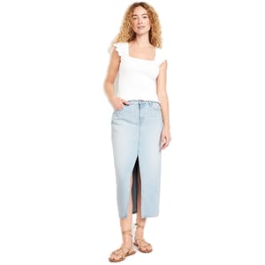 A woman is wearing a white square-neck top with ruffled sleeves, a light blue denim midi skirt with a front slit, and tan strappy sandals.