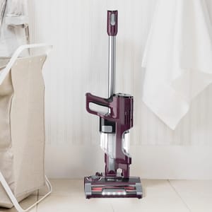 Cordless upright vacuum cleaner standing against a white bathroom wall near a hanging towel.