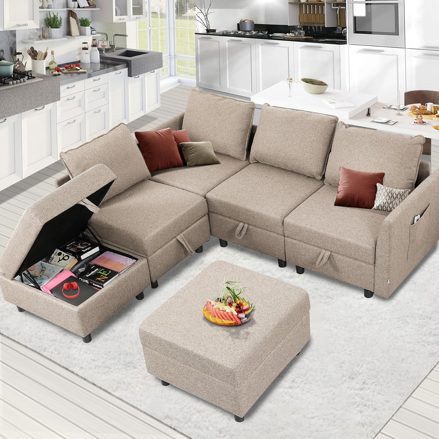 A beige modular sectional sofa with hidden storage in one seat and an ottoman topped with fruit. The living room includes a modern kitchen in the background.