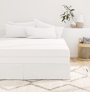 A neatly made bed with white bedding and multiple pillows, flanked by decorative vases and a plant, with a textured rug below and a woven basket to the side.