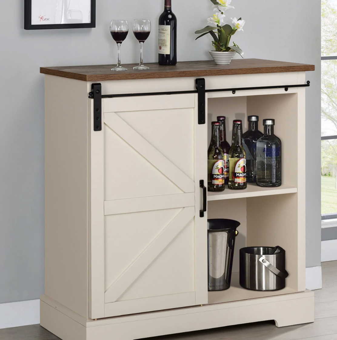 A compact sideboard featuring a sliding barn-style door concealing storage. The left side hides additional space, while the right open shelves display two cider bottles, two liquor bottles, an ice bucket, a pitcher, and a plant.