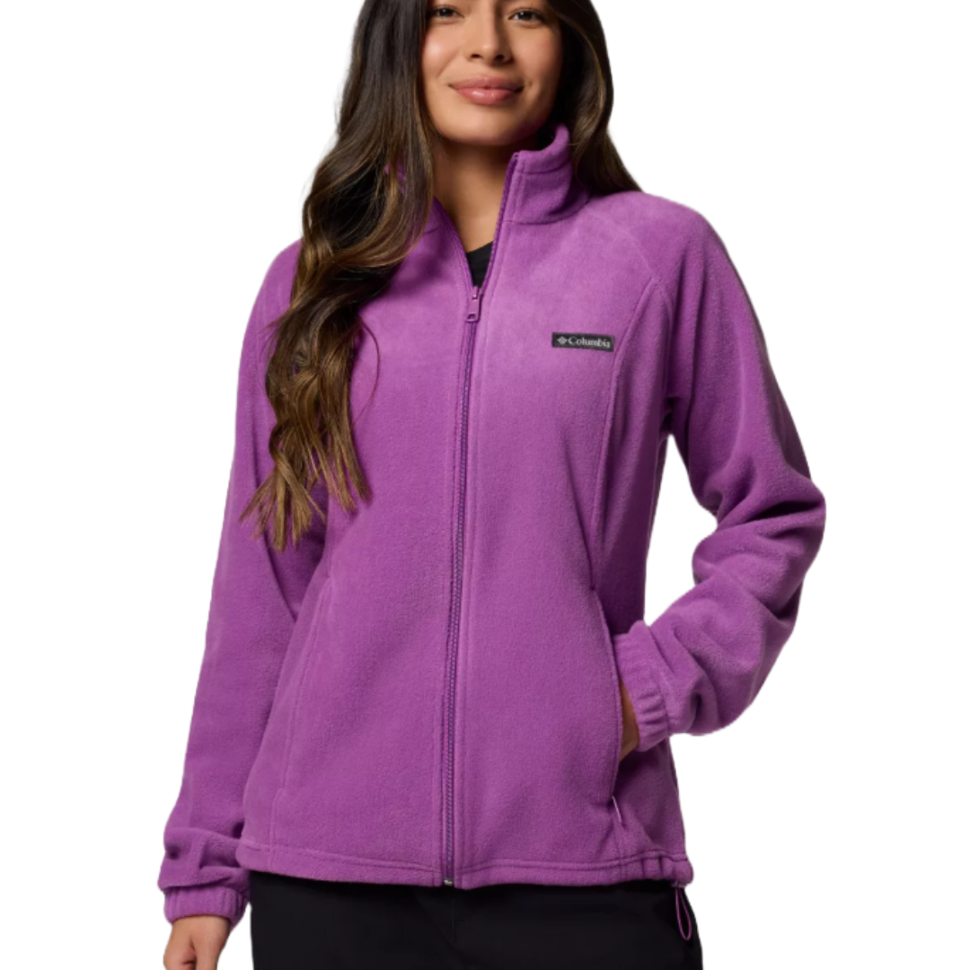 A woman wears a purple Columbia fleece jacket with a full zip and logo on the chest. The jacket includes elastic cuffs and a stand-up collar.