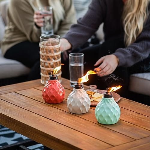 Three textured mini tabletop torches in red, white, and green are lit on a wooden table, with people in the background holding drinks.