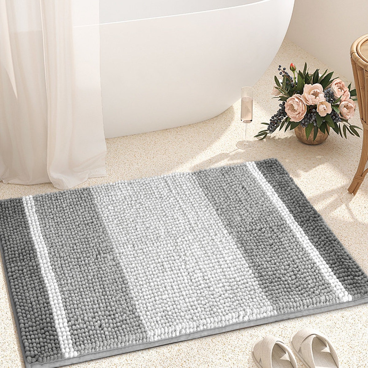 A gray and white striped bath mat is placed on a bathroom floor next to a bathtub. Nearby are a pair of slippers and a flower arrangement.