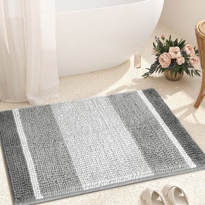 A gray and white striped bath mat is placed on a bathroom floor next to a bathtub. Nearby are a pair of slippers and a flower arrangement.
