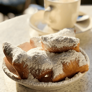 A plate of beignets dusted with powdered sugar is placed on a table beside a cup of coffee.