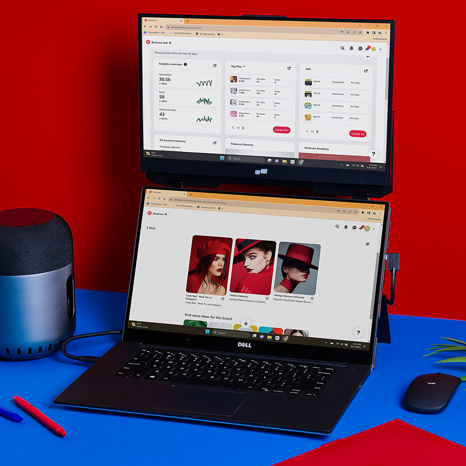 A laptop and a secondary portable screen are set up side by side on a blue and red desk with a smart speaker and wireless mouse nearby.