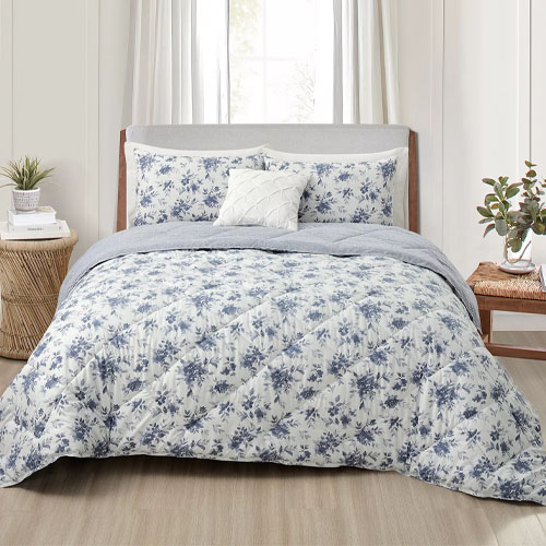 A bed with a floral-patterned comforter set, including matching pillow shams and a decorative pillow. The room features a light, neutral color scheme with a wooden headboard and natural elements like plants and a woven side table.