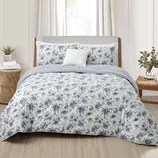 A bed with a floral-patterned comforter set, including matching pillow shams and a decorative pillow. The room features a light, neutral color scheme with a wooden headboard and natural elements like plants and a woven side table.