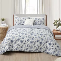 A bed with a floral-patterned comforter set, including matching pillow shams and a decorative pillow. The room features a light, neutral color scheme with a wooden headboard and natural elements like plants and a woven side table.