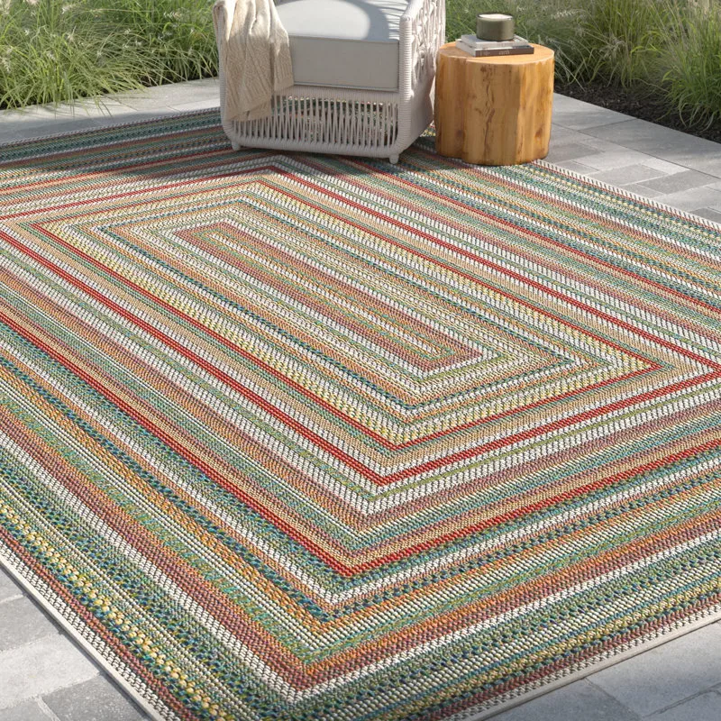 Outdoor area rug with colorful striped pattern, placed on a patio next to a wicker chair and wooden side table.