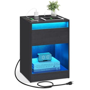 A black nightstand with integrated LED lighting features two charging ports on top for devices, a decorative plant, and an illuminated lower shelf holding an alarm clock and books.