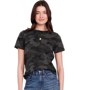 A woman models a dark camouflage-patterned t-shirt paired with high-waisted blue jeans, accessorized with a simple necklace with a small rectangular pendant.