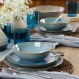 A table setting featuring light blue dishes with brown edges, blue glass tumblers, silverware, woven placemats, and light blue napkins. Cream-colored vases with small white flowers complete the arrangement.