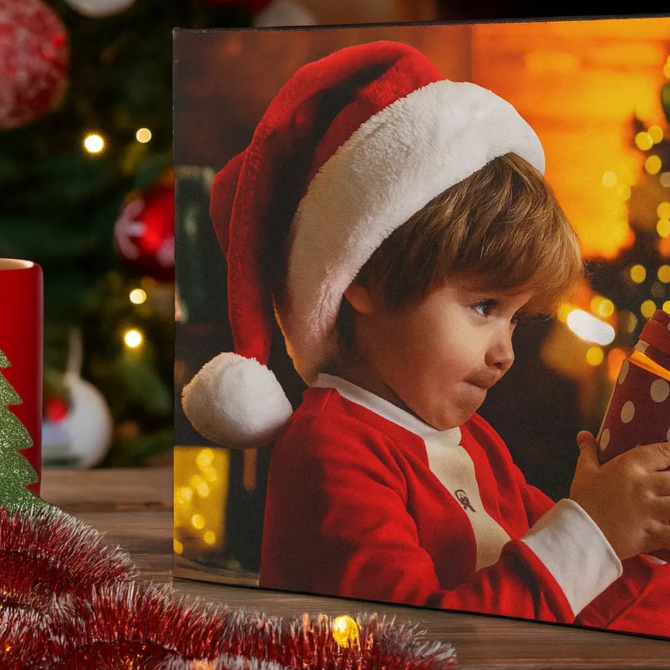 A festive scene with a canvas print of a child in a Santa hat holding a polka dot gift, next to a red mug featuring a green Christmas tree design, surrounded by tinsel and cozy holiday lights.