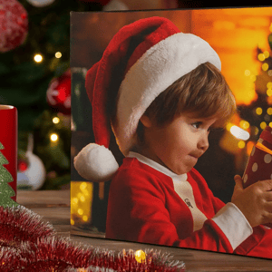 A festive scene with a canvas print of a child in a Santa hat holding a polka dot gift, next to a red mug featuring a green Christmas tree design, surrounded by tinsel and cozy holiday lights.