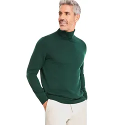 A man is wearing a dark green turtleneck sweater paired with light-colored trousers, standing with his hand in his pocket.