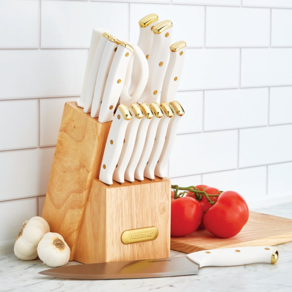 Knife block set with white handles and gold accents; includes various knives on a wooden block. In the background are tomatoes on a cutting board and garlic, placed on a marble countertop against a white tiled wall.